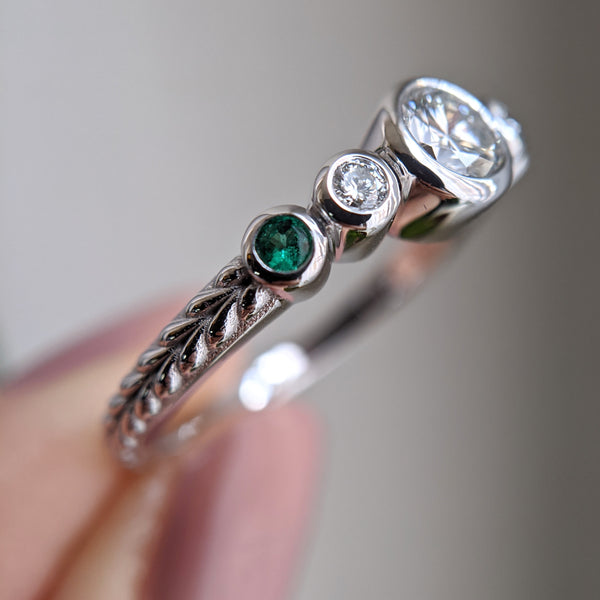 Diamond and emerald ring, stones set in bezels, with braid engraving on the shank. The ring is held by a hand with pale pink nails.