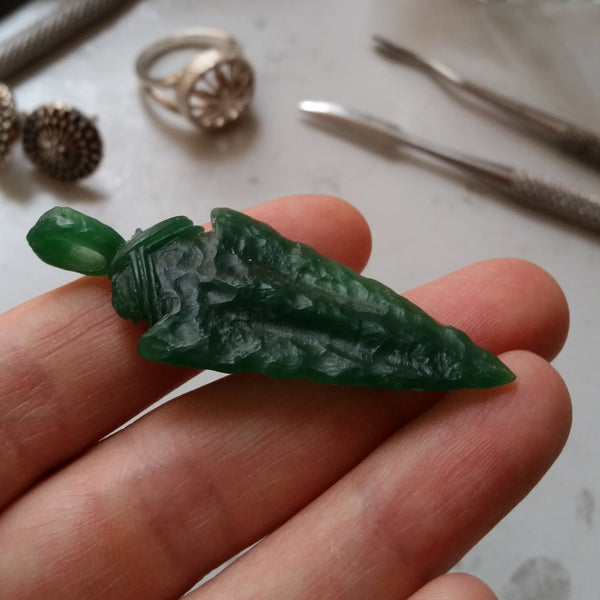 Carved green wax model of an arrowhead being held in a hand. In the background are silver rings and wax carving tools.