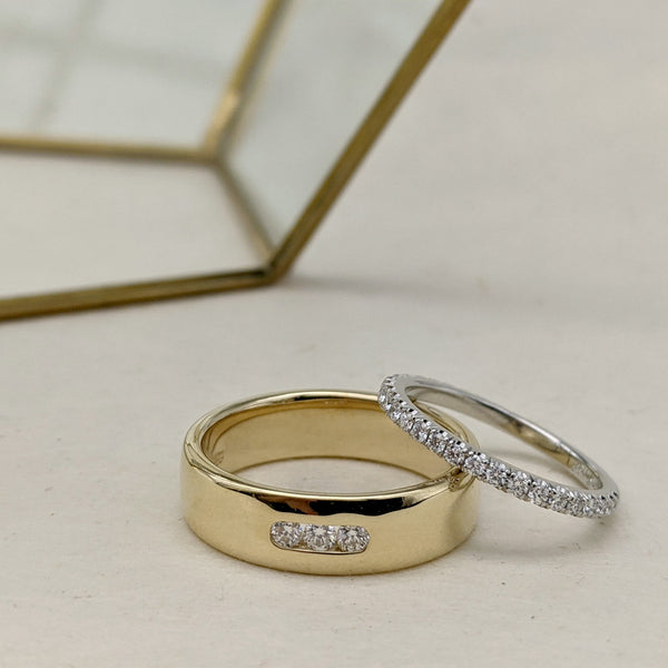 Yellow gold wedding ring with three channel set diamonds, with a platinum and diamond pavé wedding ring leaning against it. Rings are on a cream backgrop, with a geometric brass metal decoration in the background.