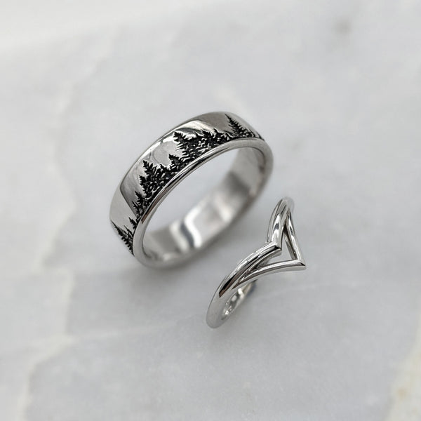 Double chevron wedding ring on the bottom, with a wide wedding ring above it. The wide ring has a blackened treeline design. Both rings are standing on a piece of gray marble.