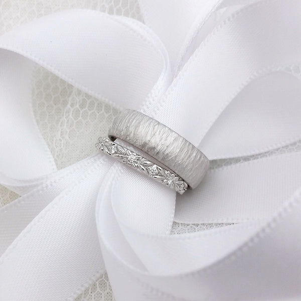 White gold and diamond eternity wedding ring with floral engraving, next to a wide wedding ring with a hand carved line texture. Both rings have white ribbon threaded through them.