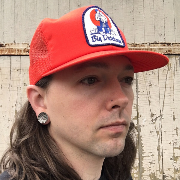 White male with long, dark blonde hair and a red trucker hat wearing a pair of sterling silver gauge earrings with a botanical engraved pattern.