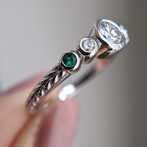 Diamond and emerald ring, stones set in bezels, with braid engraving on the shank. The ring is held by a hand with pale pink nails. 