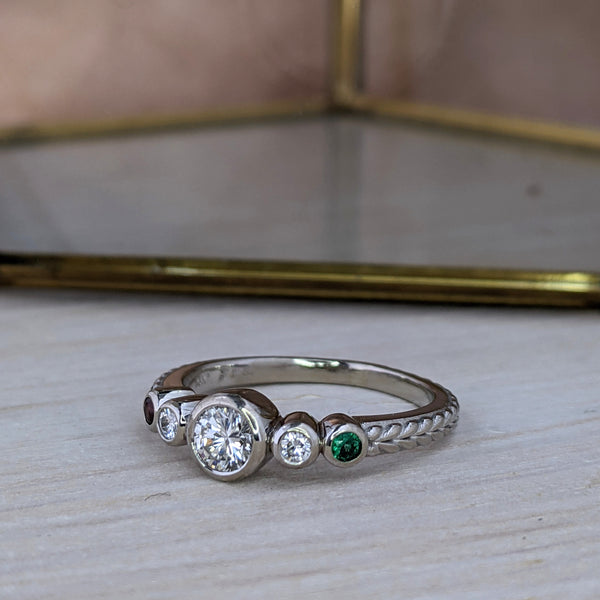 Diamond and emerald ring, stones set in bezels, with braid engraving on the shank. The ring is on pale gray wood, in front of a brass sculpture with a peach background.