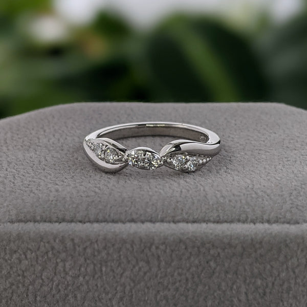 White gold and diamond wedding ring on a gray suede box, with green leaves in the background.