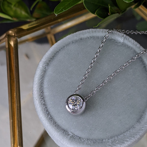 Round brilliant diamond set in a bezel setting, on a cable chain. The pendant sits atop a gray ring box lid, with green leaves along the top of the image.