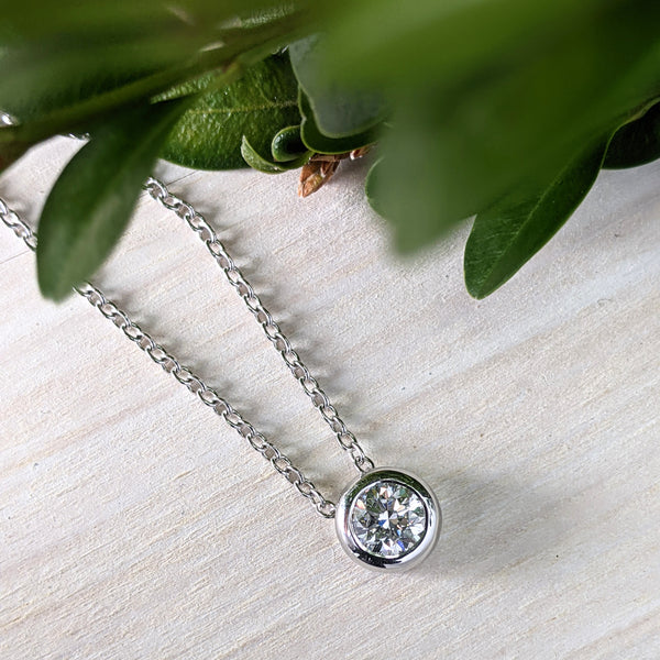 Round brilliant diamond set in a bezel setting, on a cable chain. The pendant sits atop a pine background, with green leaves along the top of the image.